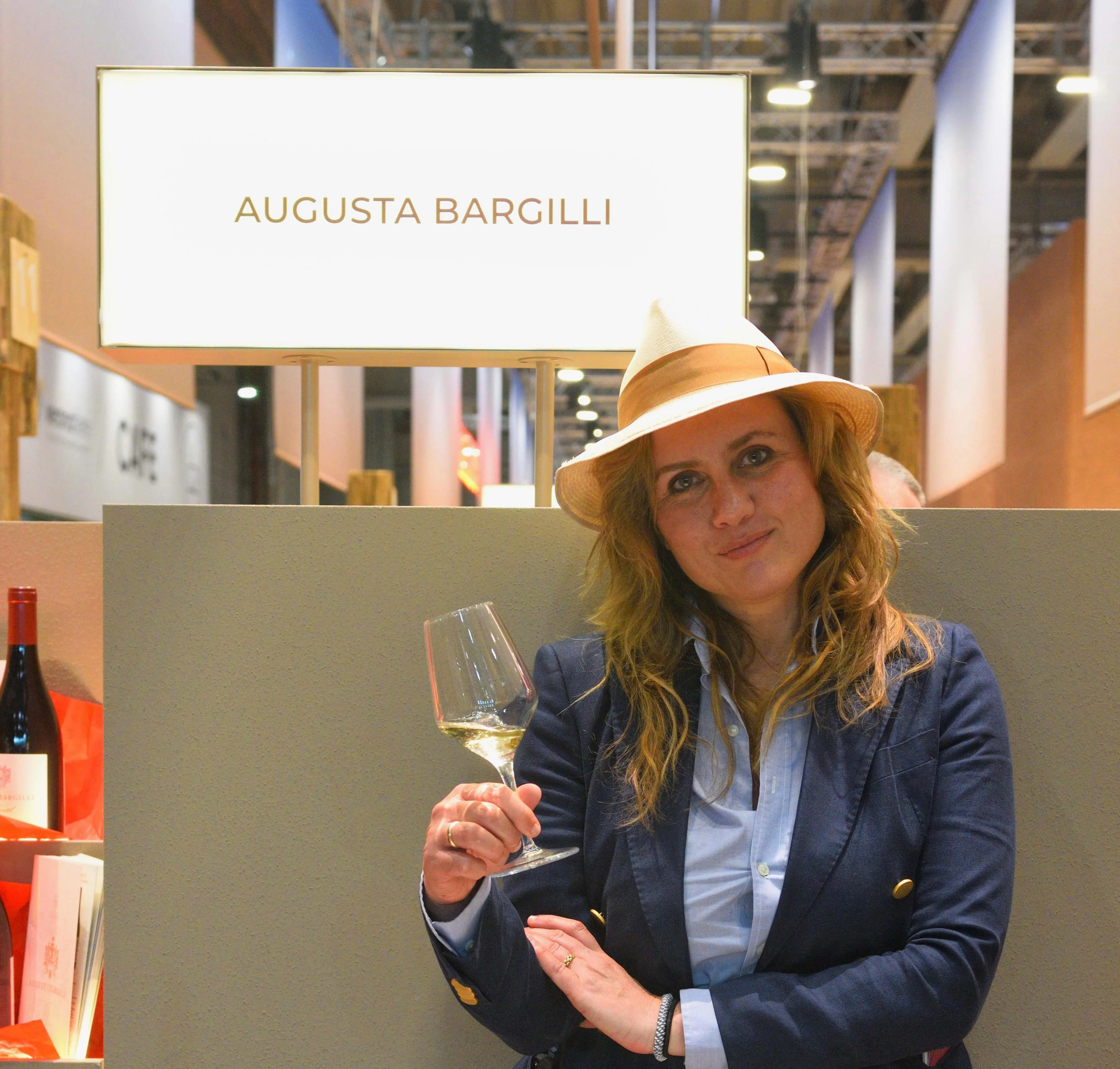 Augusta Bargilli al suo stand in fiera, con il cappello di paglia e un calice di bianco, sotto l’insegna con il suo nome