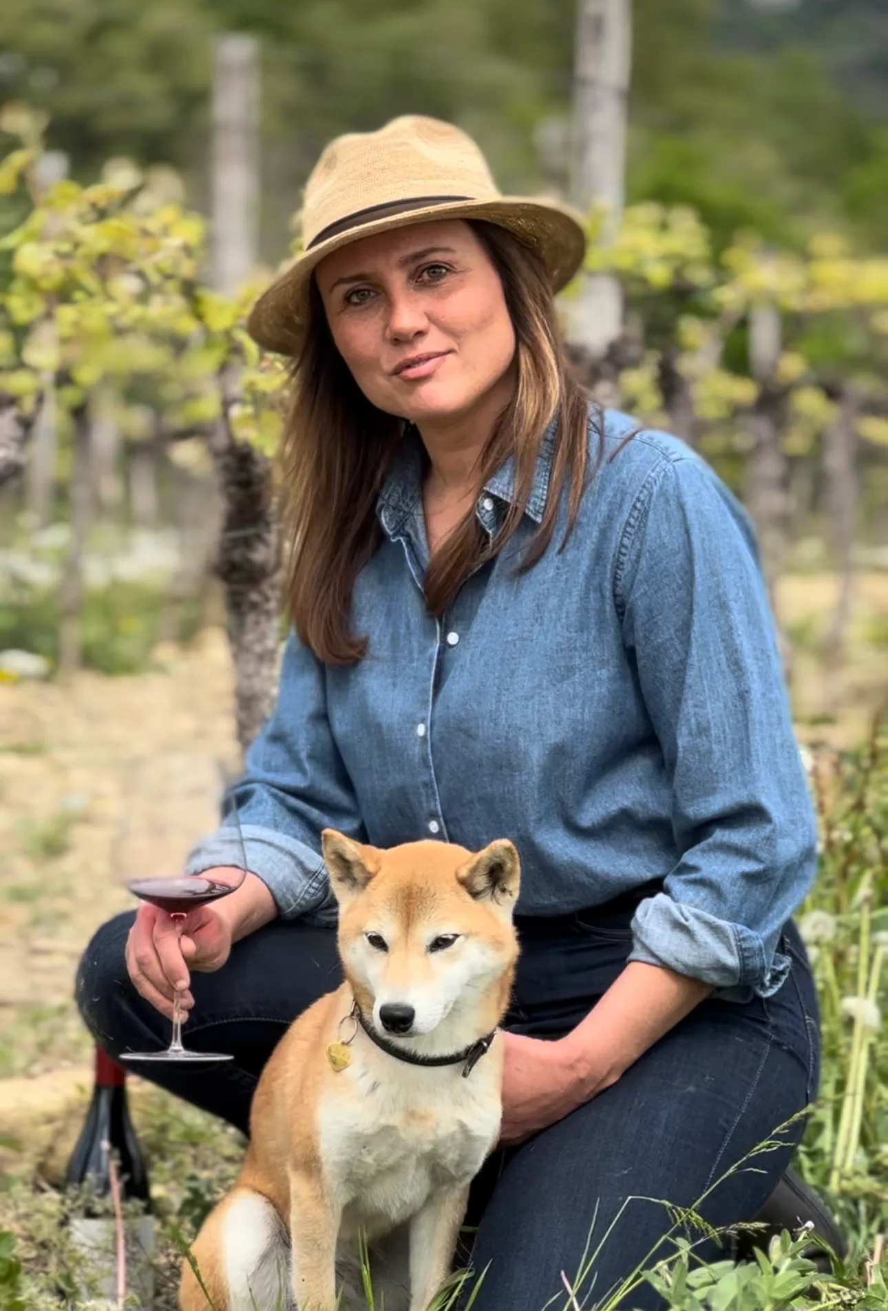 Augusta Bargilli inginocchiata in vigna con il cappello di paglia, un calice di rosso in mano, accanto al suo cane