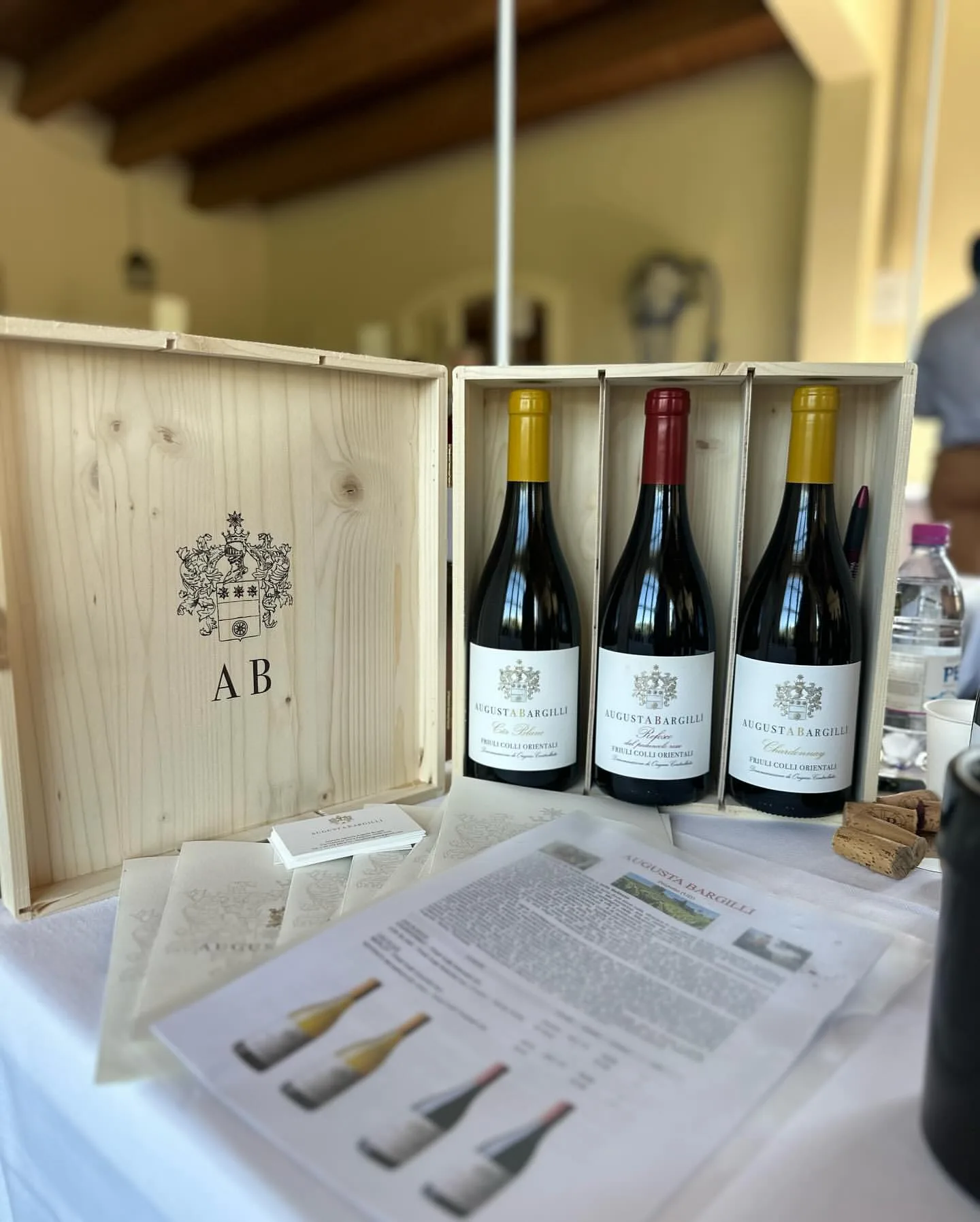 Una cassa di legno con lo stemma e le lettere AB, aperta su tre bottiglie, accanto alle schede dei vini