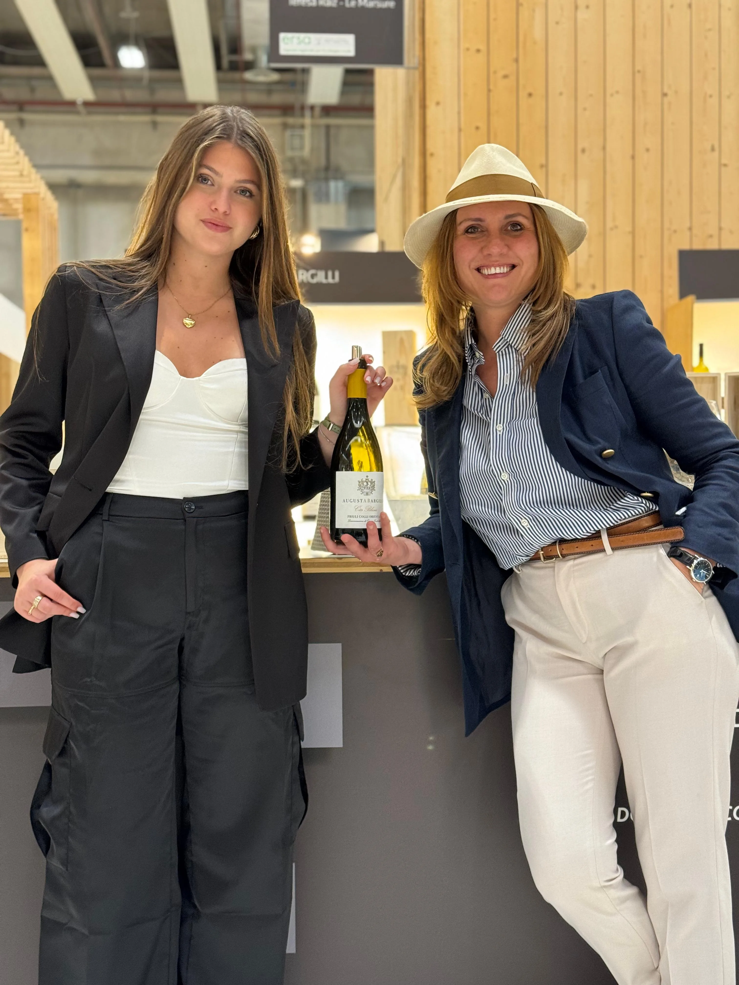 Augusta Bargilli e la figlia Aloisa al loro stand al Vinitaly: Aloisa in blazer nero tiene una bottiglia, Augusta, con il cappello di paglia, si appoggia al banco